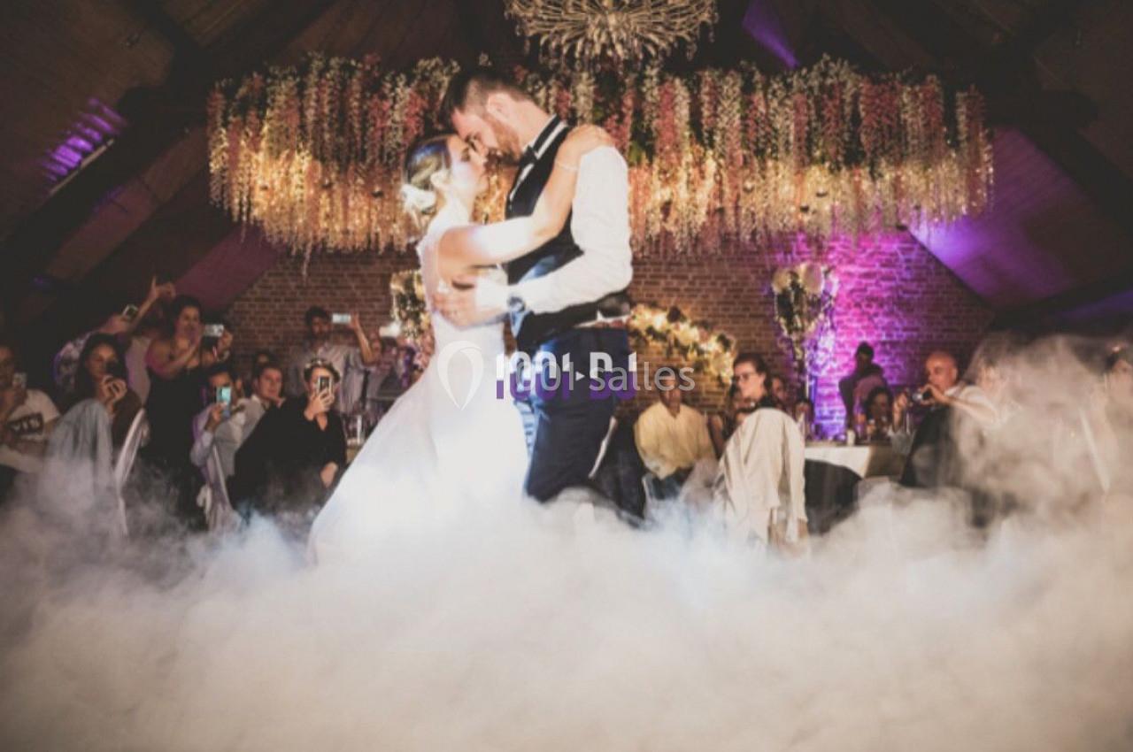 Un couple danse sous des lumières colorées et une décoration florale, entouré de fumée et d'invités.