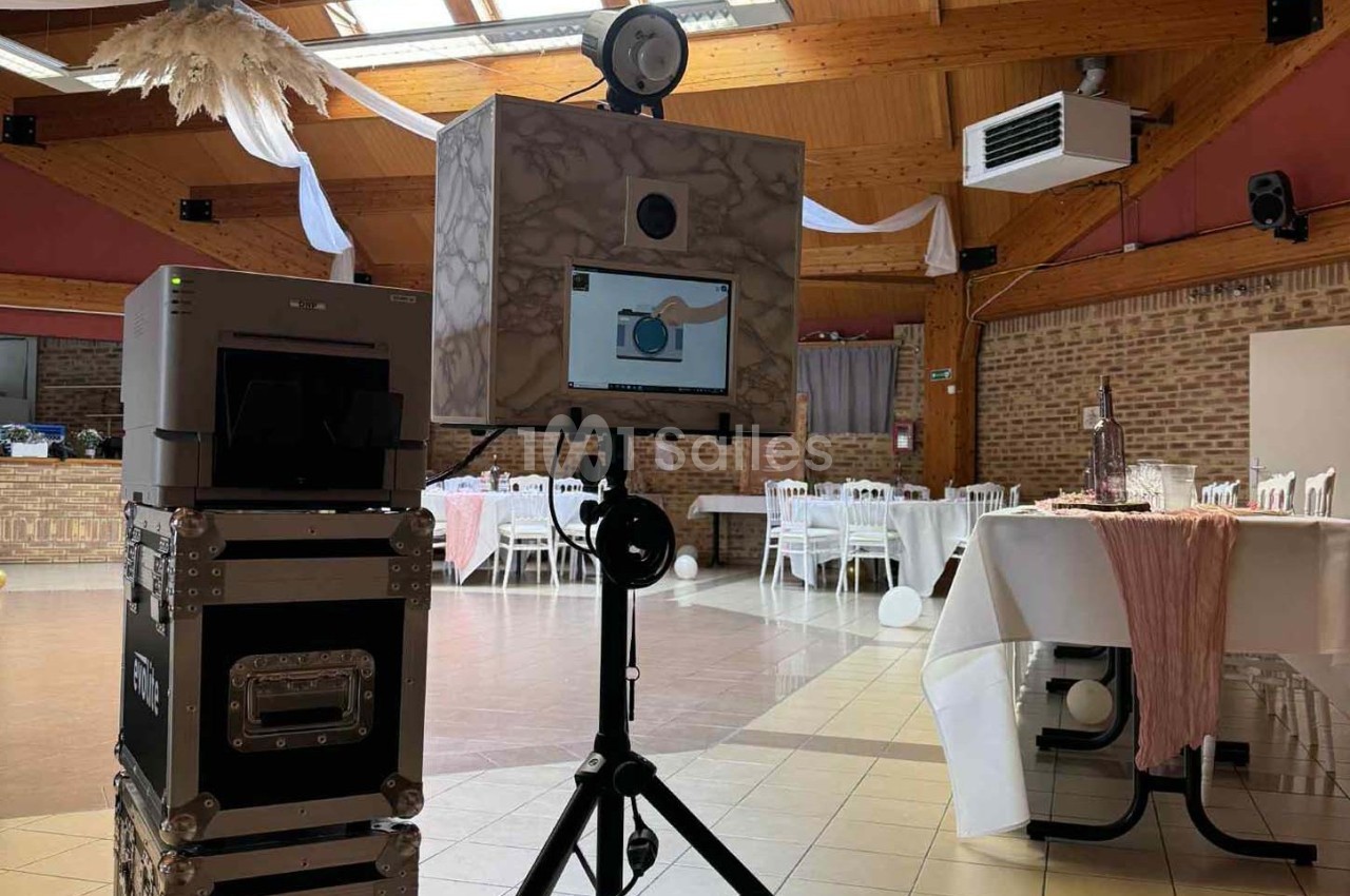 Borne photo installée dans une salle de réception décorée, avec tables dressées et éléments suspendus au plafond.