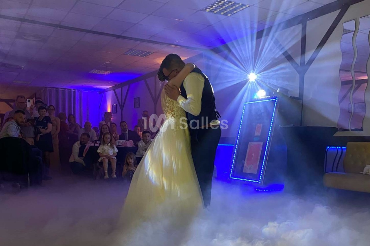 Un couple danse enlacé sous des lumières tamisées, entouré de spectateurs dans une salle décorée.