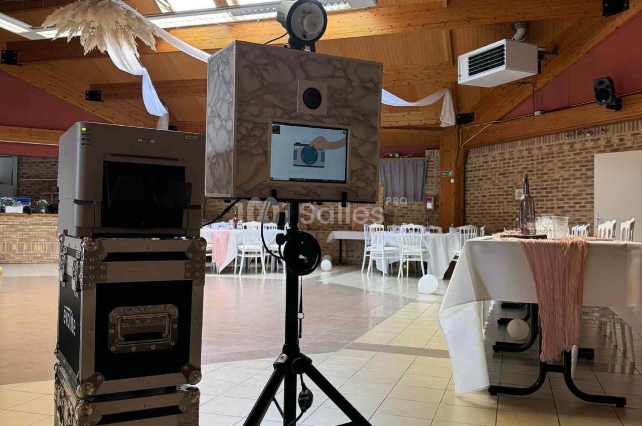 Borne photo installée dans une salle de réception avec tables décorées et lumière naturelle entrant par le toit.