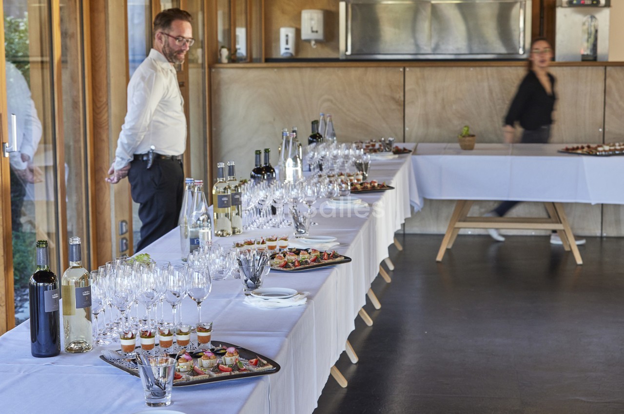 Table dressée avec verres, bouteilles et plateaux de canapés dans une salle lumineuse, un homme et une femme en arrière-plan.