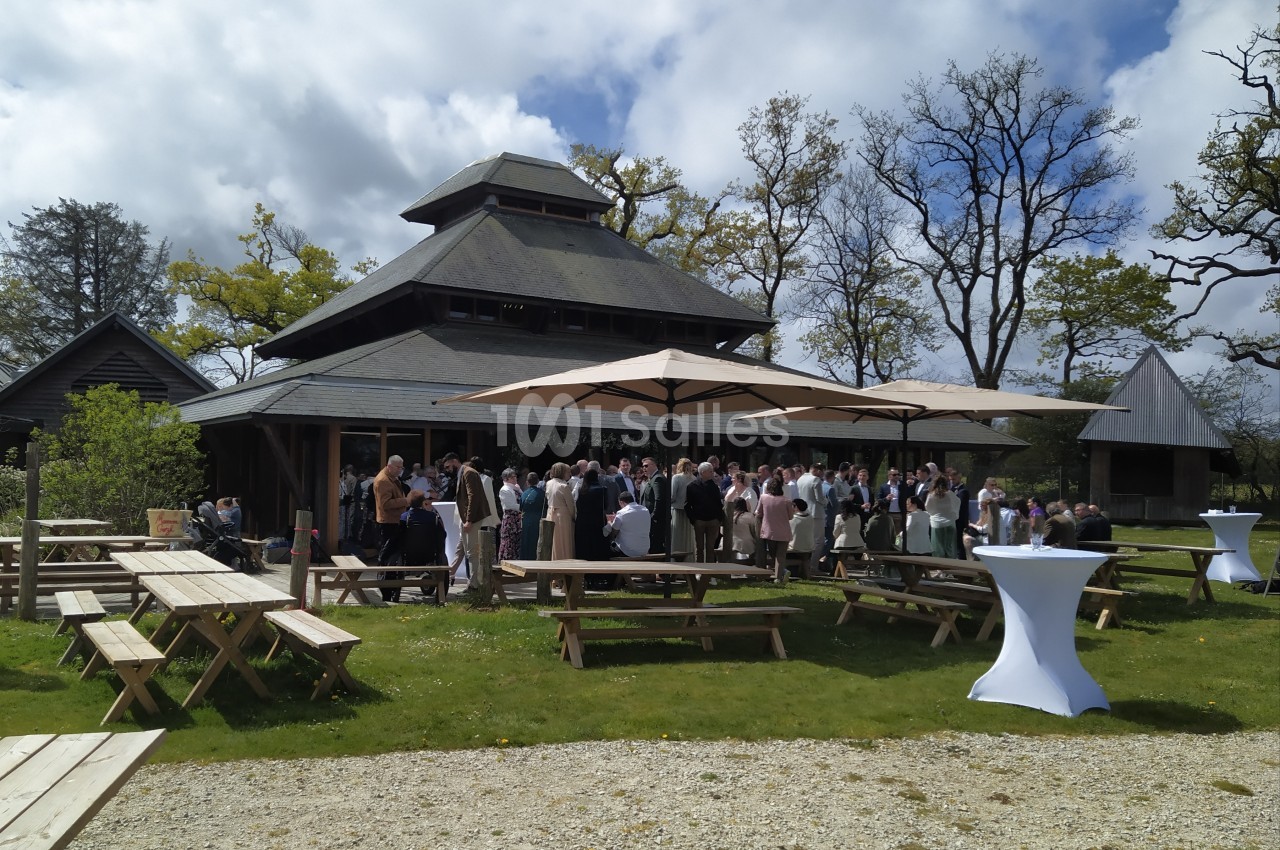 Groupe de personnes rassemblées devant un bâtiment en bois avec des tables de pique-nique et des parasols dans un jardin.