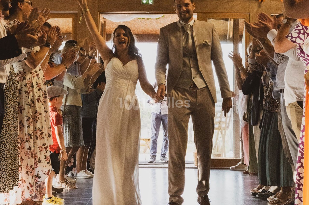 Un couple de mariés souriant entre dans une salle, entouré d'invités applaudissant.