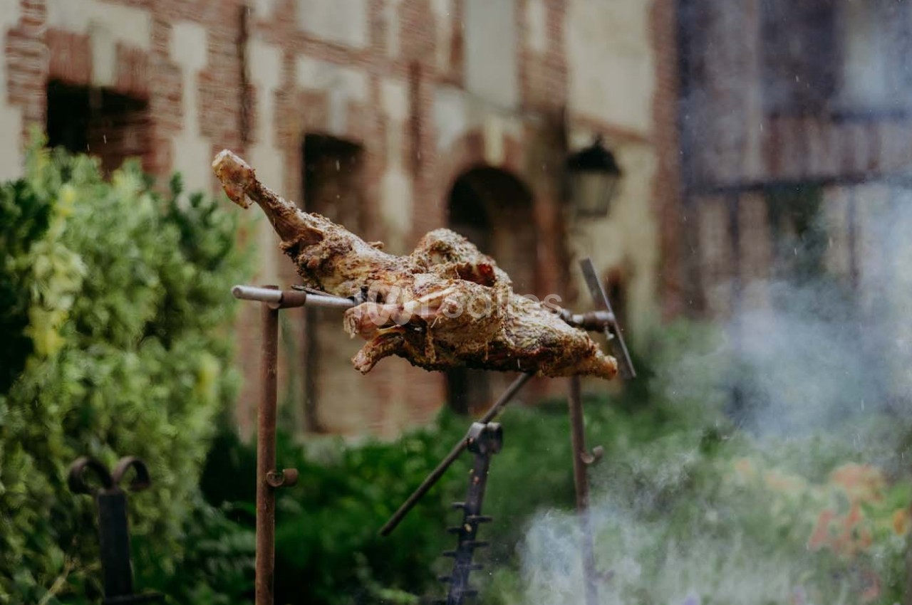 Viande rôtie sur une broche en extérieur, devant un mur en briques et un jardin verdoyant.
