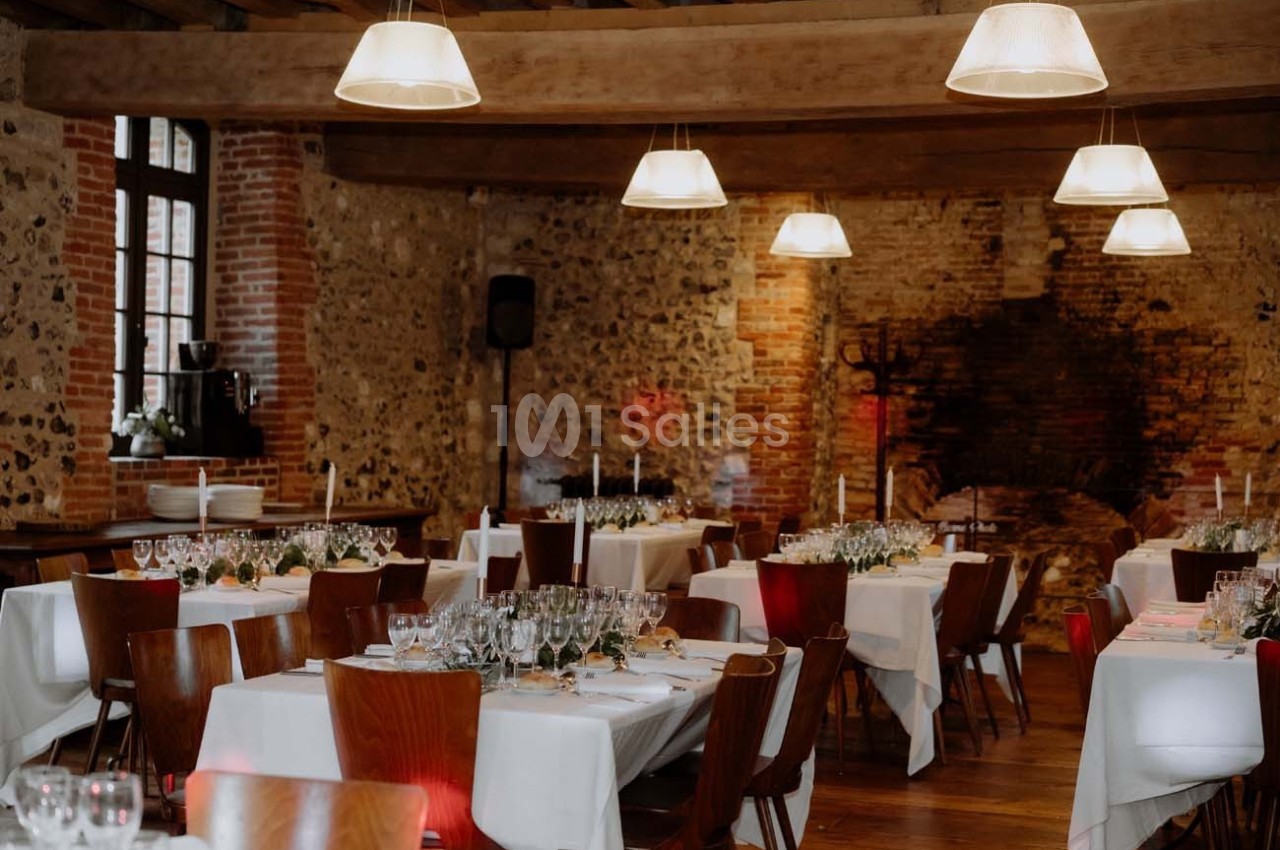 Salle de réception avec murs en pierre, tables dressées avec nappes blanches, chaises en bois et éclairage suspendu.