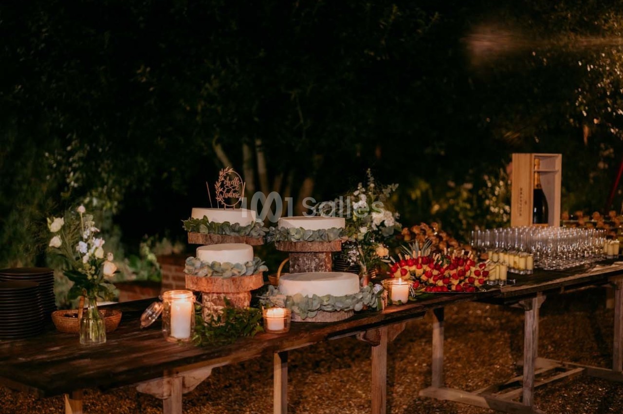 Table en bois décorée pour un événement, avec gâteaux, bougies, fleurs et verres disposés sous un éclairage tamisé.