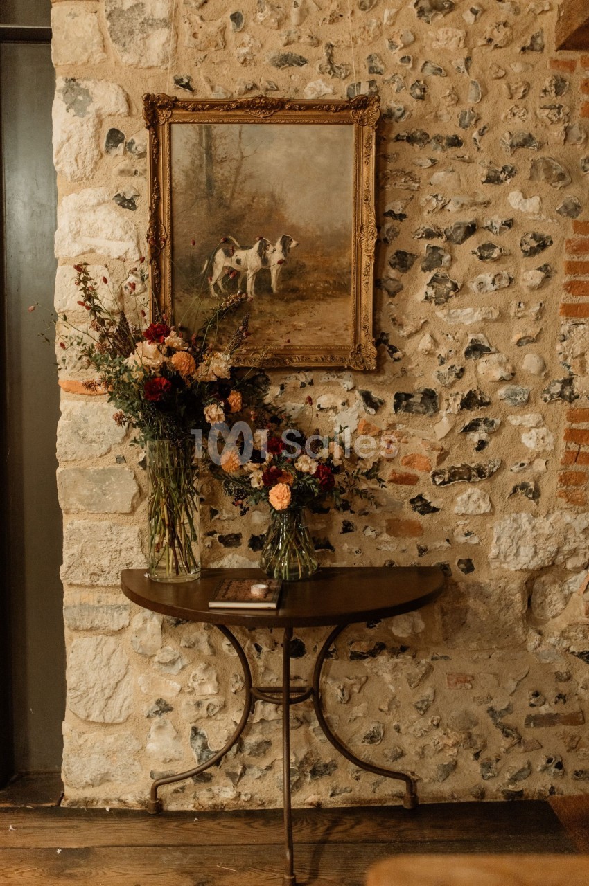 Table en bois avec deux bouquets de fleurs devant un mur en pierre, surmontée d'un tableau représentant des chiens.