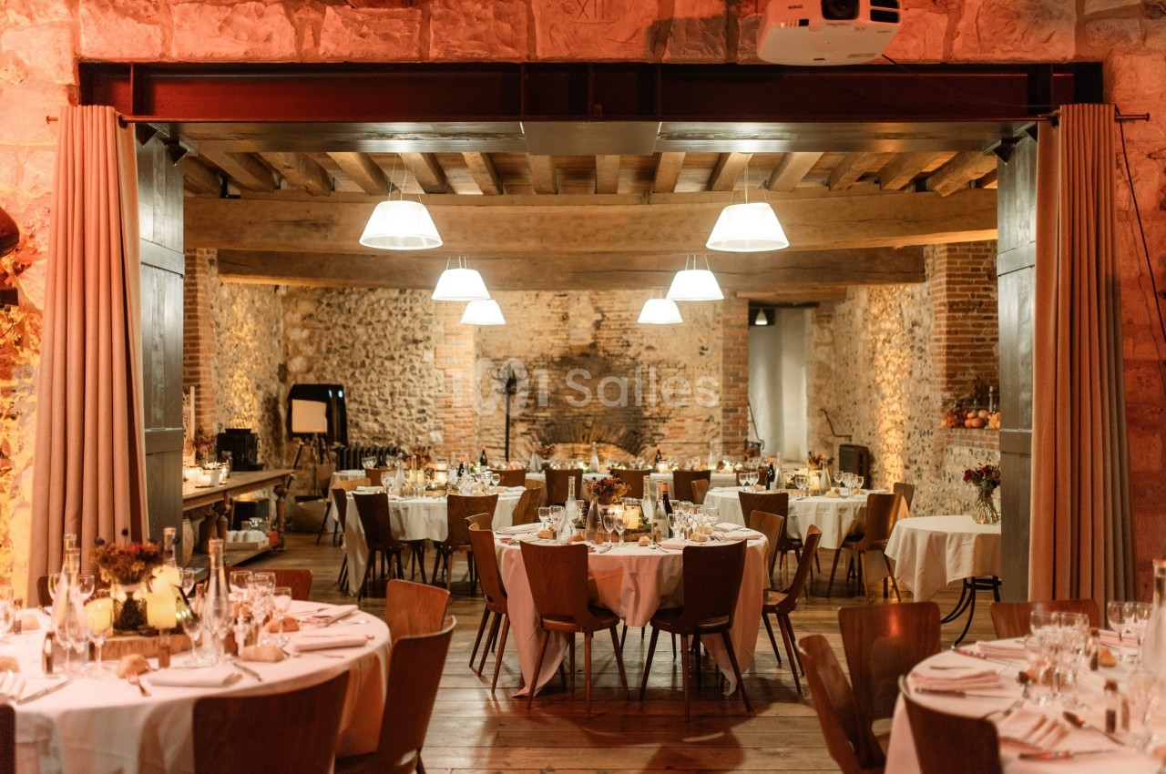 Salle de réception chaleureuse avec tables rondes dressées, éclairage doux et murs en pierre apparente.