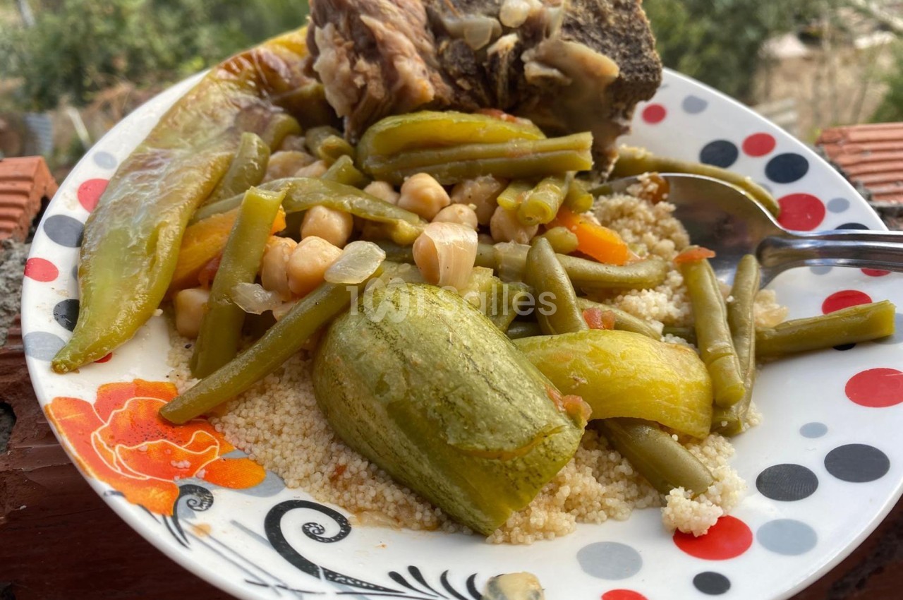 Assiette de couscous avec légumes variés, pois chiches, haricots verts et morceau de viande, servie en extérieur.
