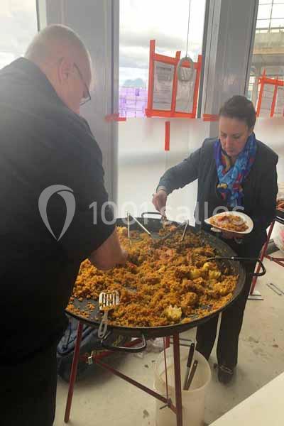 Deux personnes se servent de la paella dans une grande poêle lors d'un événement en intérieur.