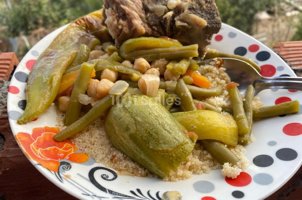 Assiette de couscous avec légumes variés, pois chiches, haricots verts et morceau de viande, servie en extérieur.