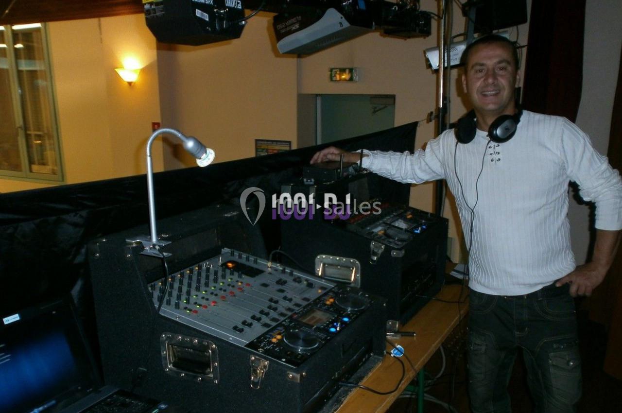 Un homme souriant se tient près d'une table de mixage et d'équipements audio dans une salle éclairée.