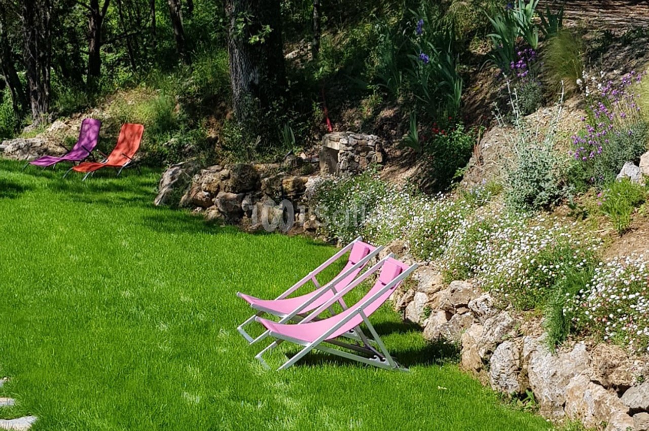 Deux chaises longues roses sur une pelouse verte, bordées de fleurs et d'arbustes sous des arbres.