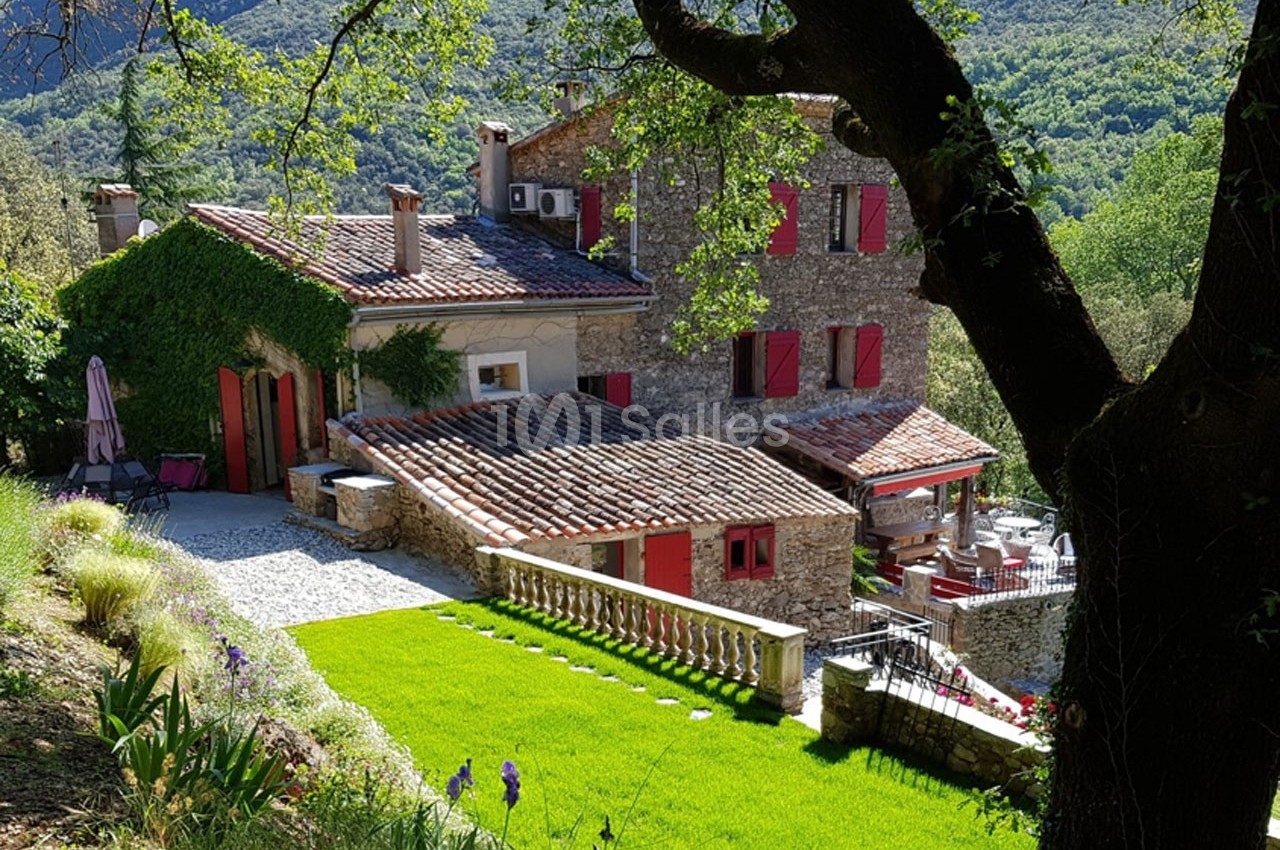 Maison en pierre avec volets rouges entourée de verdure et située dans un paysage vallonné et boisé.