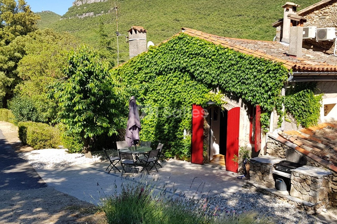 Maison en pierre avec façade recouverte de lierre, volets rouges et terrasse aménagée, entourée de verdure.