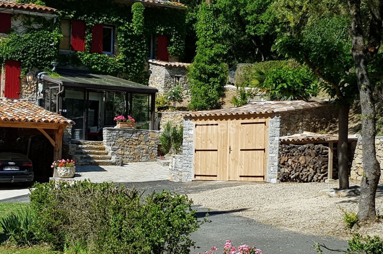 Maison en pierre avec volets rouges entourée de végétation, garage en bois et abri pour le bois de chauffage.