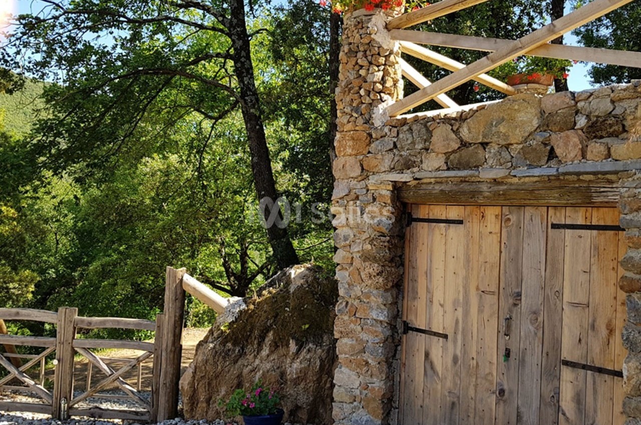 Façade en pierre avec une porte en bois, terrasse fleurie au-dessus, entourée de végétation et d'une clôture en bois.