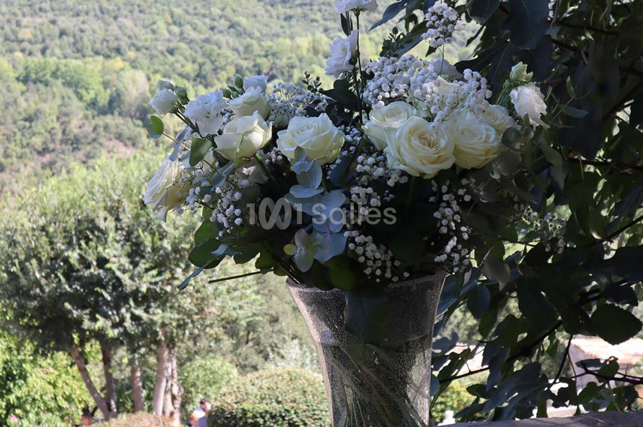 Bouquet de fleurs blanches et vertes dans un vase en verre, posé devant un paysage boisé en arrière-plan.