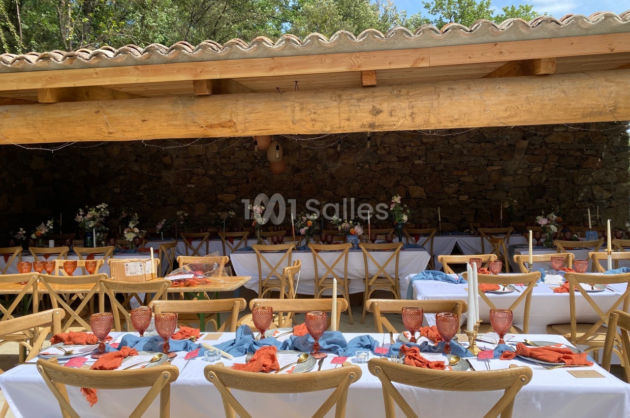 Tables décorées avec nappes blanches, serviettes colorées et vaisselle en terre cuite sous une pergola en bois.