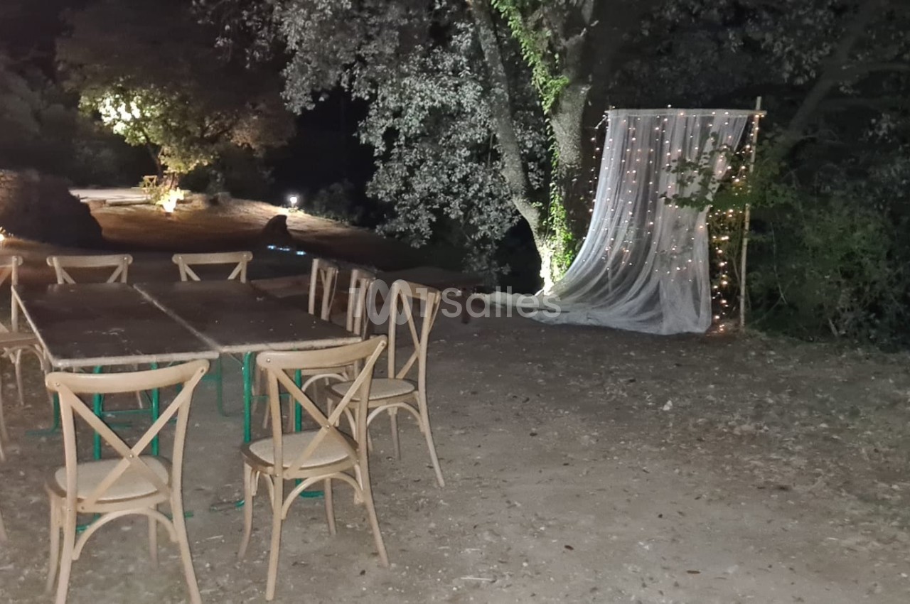 Chaises en bois disposées autour de tables en extérieur, avec un décor lumineux et un rideau blanc sous des arbres la nuit.