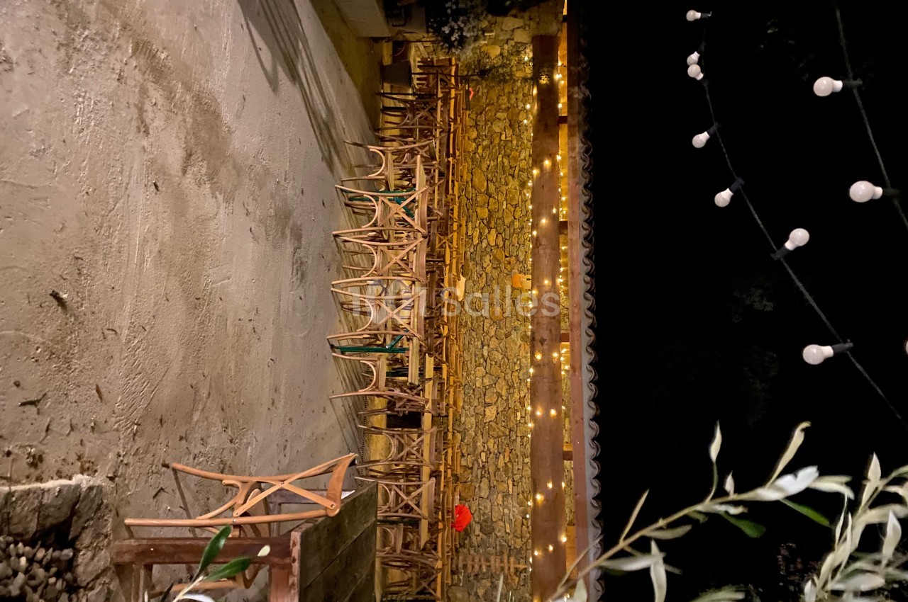 Terrasse extérieure éclairée par des guirlandes lumineuses, avec des chaises en bois alignées contre un mur en pierre.