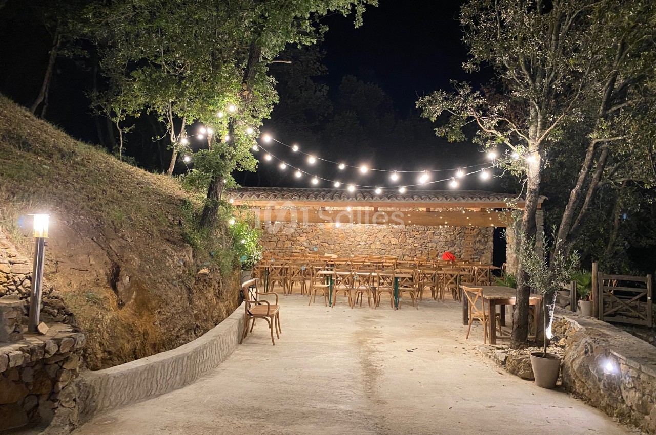 Terrasse extérieure éclairée par des guirlandes lumineuses, avec tables et chaises en bois, entourée de végétation.