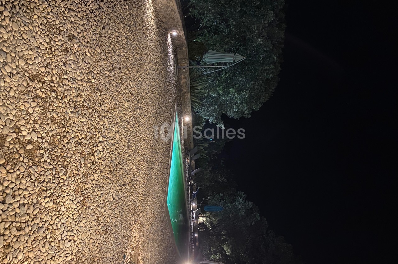 Piscine éclairée entourée de galets et d'arbres dans un jardin, photographiée de nuit.