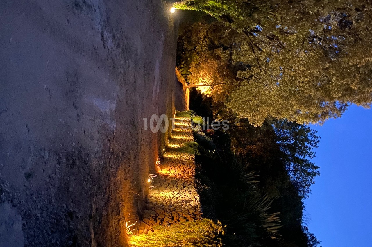 Allée bordée de murs en pierre et de végétation éclairée par des lumières au crépuscule.
