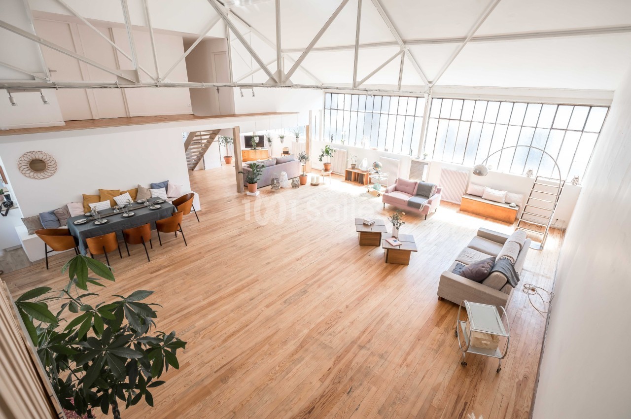 Loft spacieux avec parquet clair, grandes fenêtres, coin salon, salle à manger et mezzanine visible.
