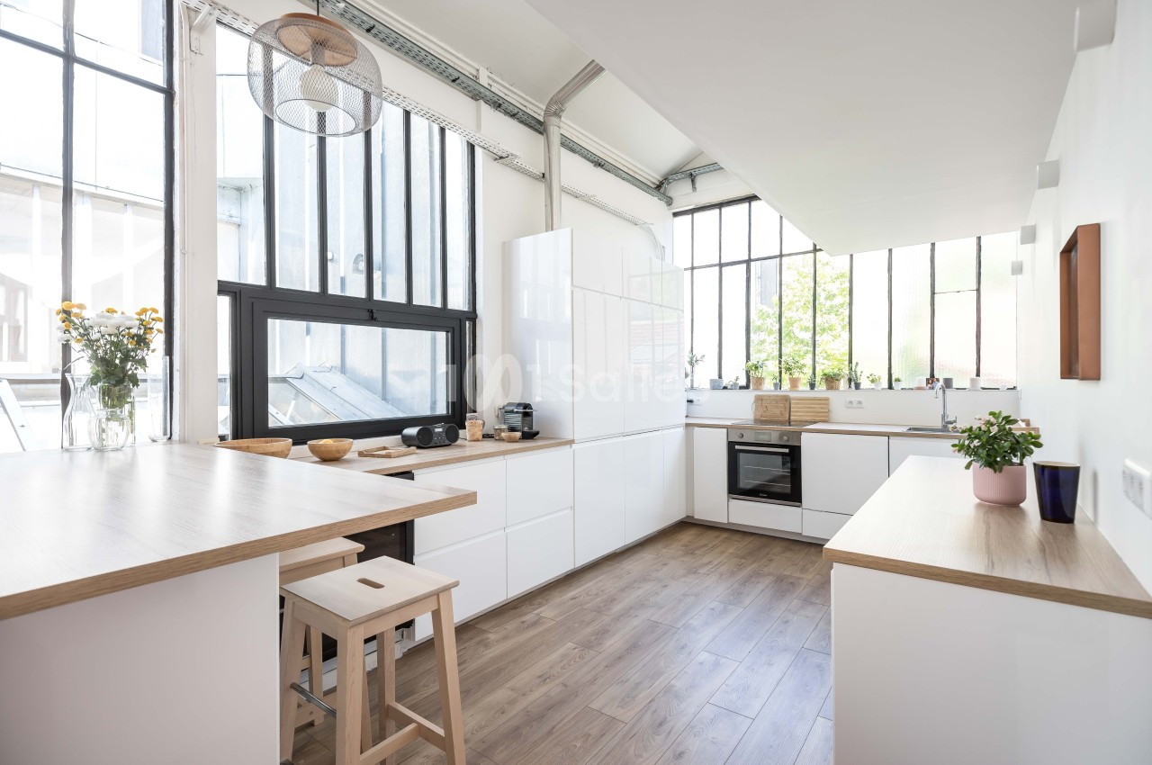 Cuisine lumineuse avec meubles blancs, plan de travail en bois, grandes fenêtres et sol en parquet clair.
