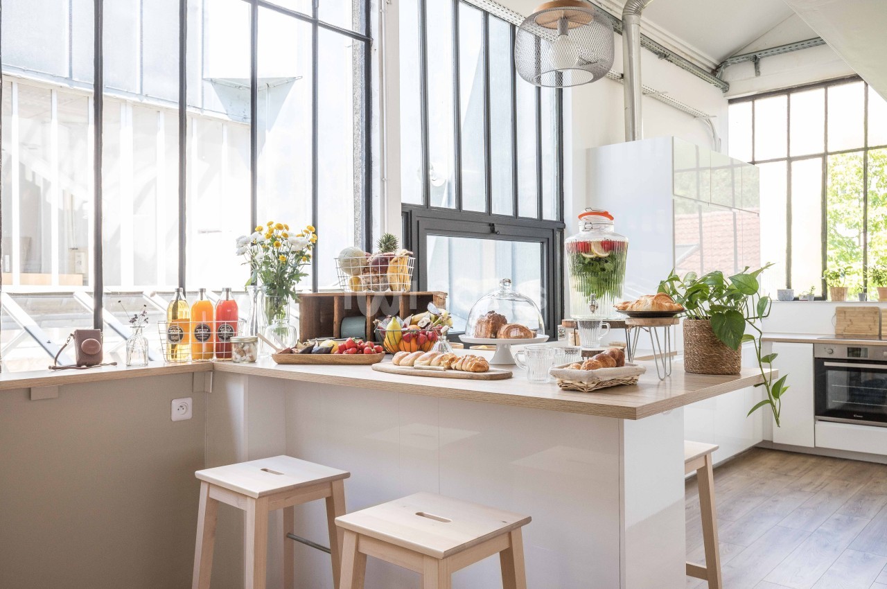 Cuisine lumineuse avec grande verrière, comptoir en bois garni de viennoiseries, fruits, jus et plantes décoratives.