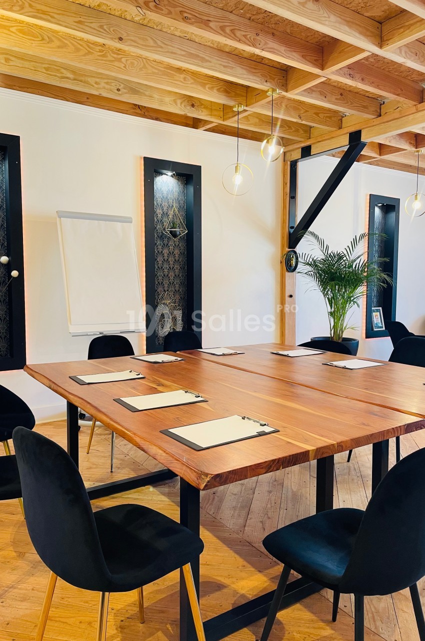 Salle de réunion lumineuse avec table en bois, chaises noires, paperboard et décoration végétale.