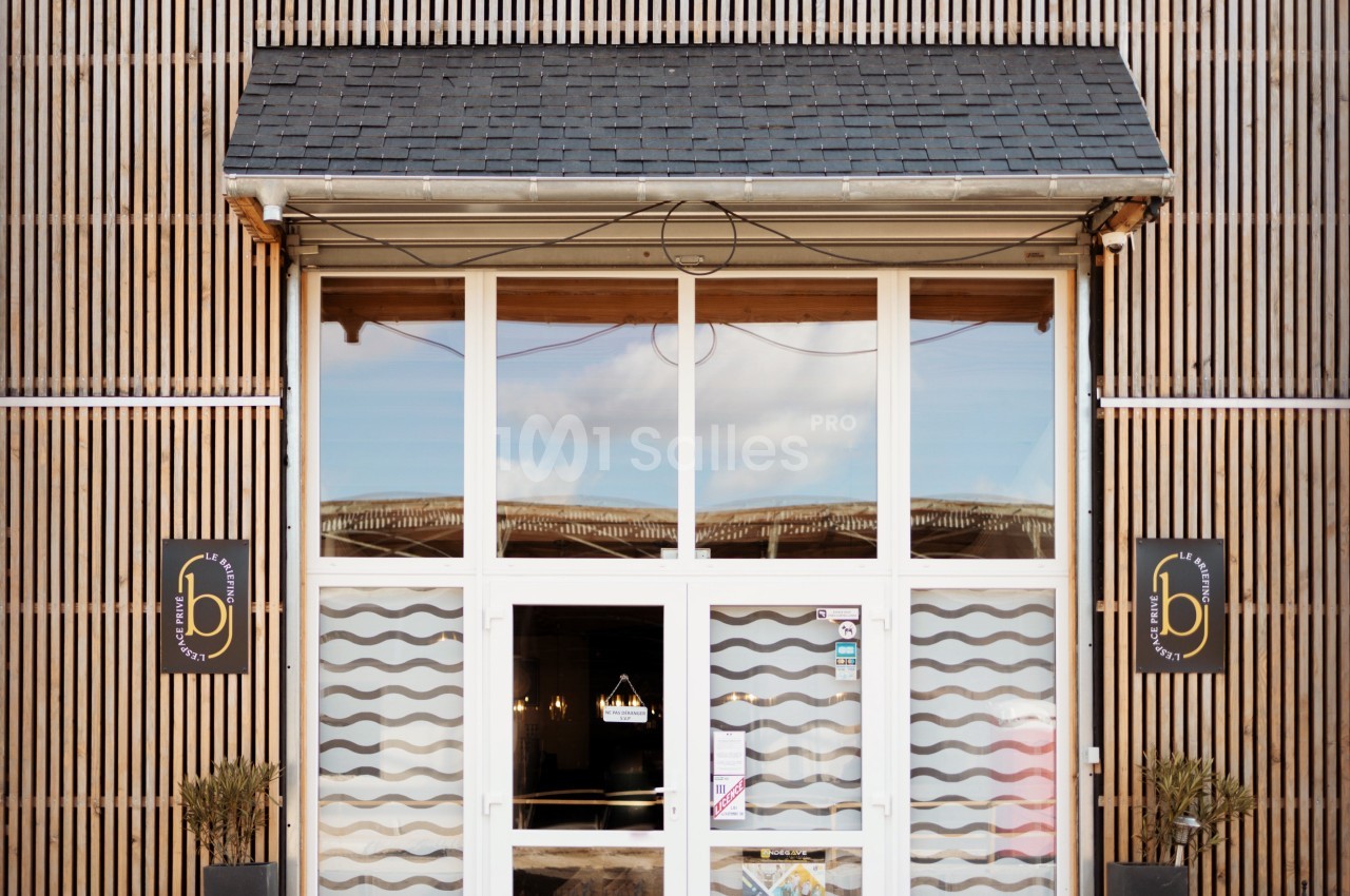 Façade d'un bâtiment avec une porte vitrée encadrée de bois et deux panneaux noirs portant un logo doré.