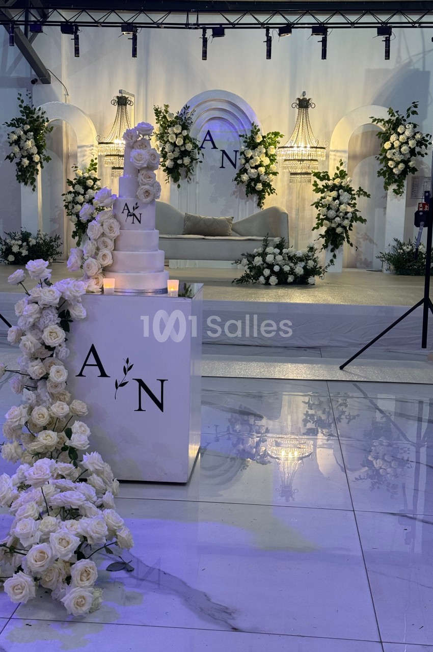 Décor de mariage élégant avec un gâteau blanc, des roses blanches et une scène ornée de fleurs et d'arches lumineuses.