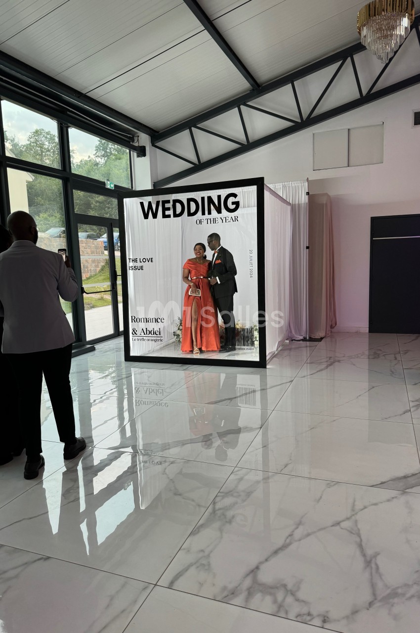 Un couple pose dans une cabine photo décorée pour un mariage, entouré de quelques invités dans une salle lumineuse.