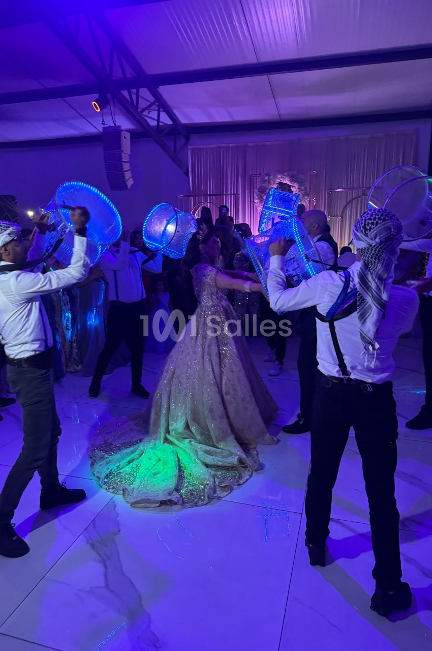 Des danseurs en tenue traditionnelle entourent une mariée sous des lumières colorées dans une salle de réception.