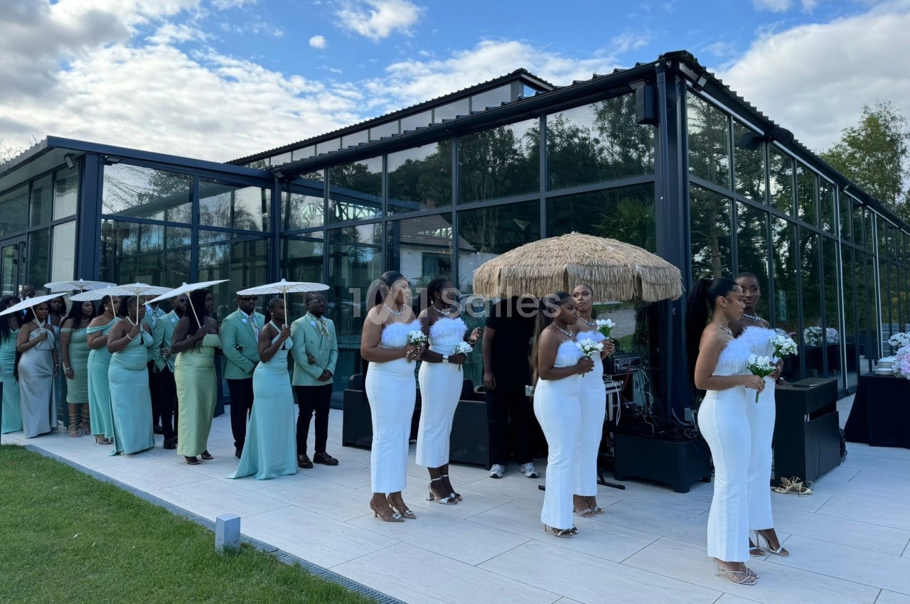 Un cortège de mariage en tenue élégante se tient devant un bâtiment vitré, sous un ciel partiellement nuageux.