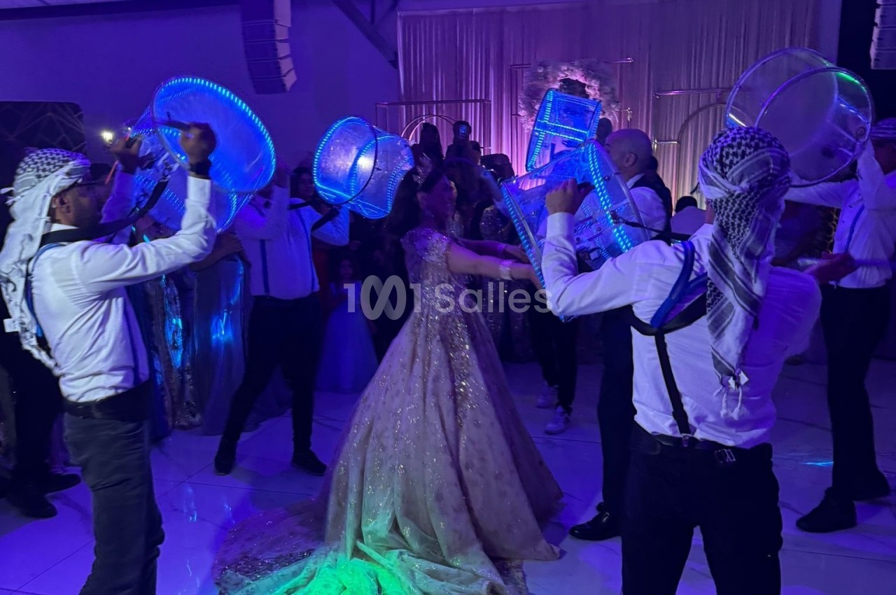 Des danseurs en tenue traditionnelle jouent du tambour lumineux autour d'une femme en robe de soirée dans une salle éclairée.