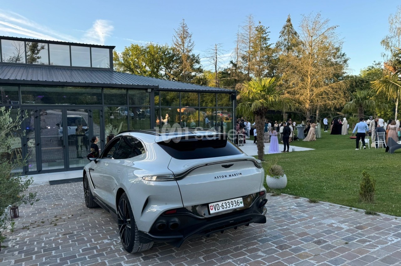 Voiture grise garée sur une allée pavée devant un bâtiment vitré, avec des invités rassemblés dans un jardin.
