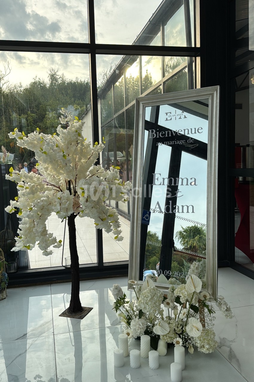 Panneau de bienvenue pour un mariage, avec un miroir encadré, des fleurs blanches et des bougies décoratives.