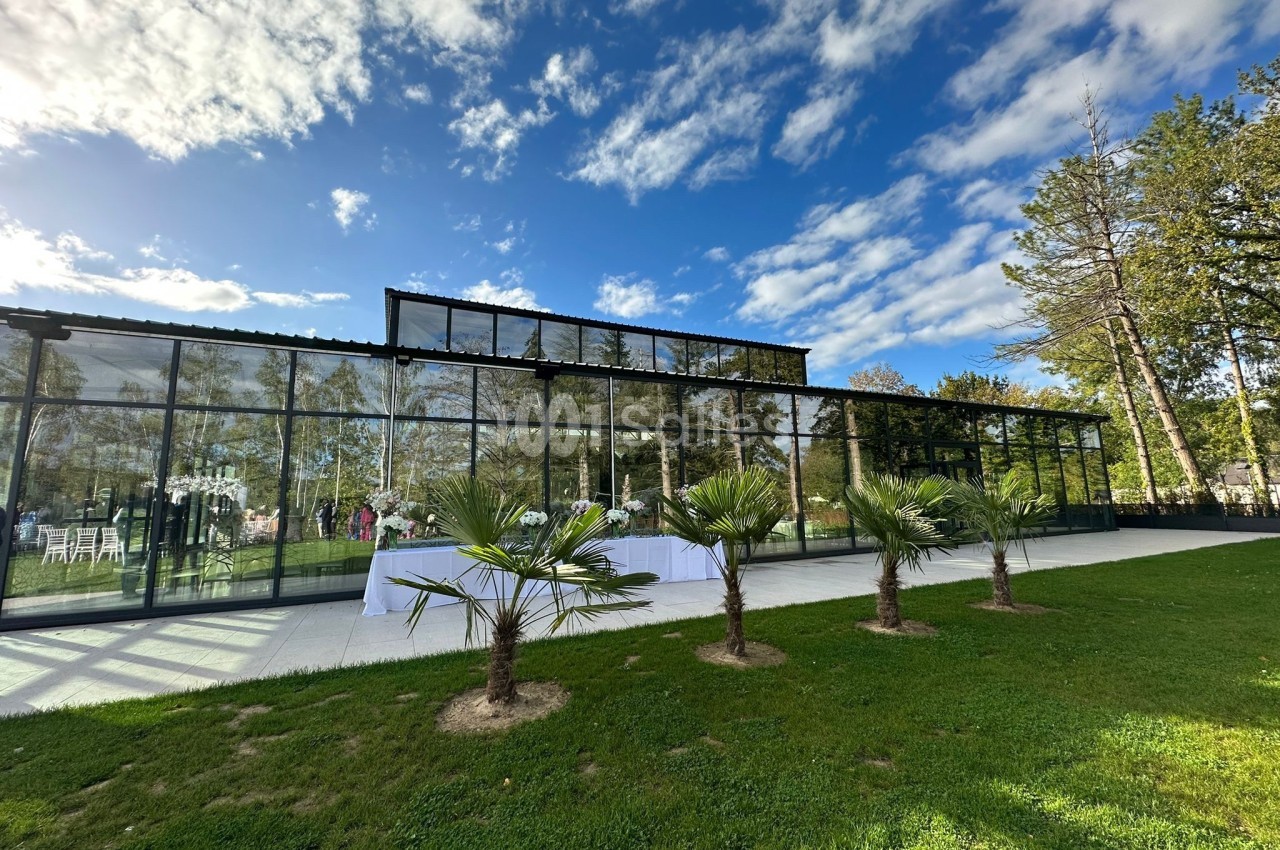 Salle vitrée moderne entourée de verdure, avec des palmiers au premier plan et un ciel partiellement nuageux.