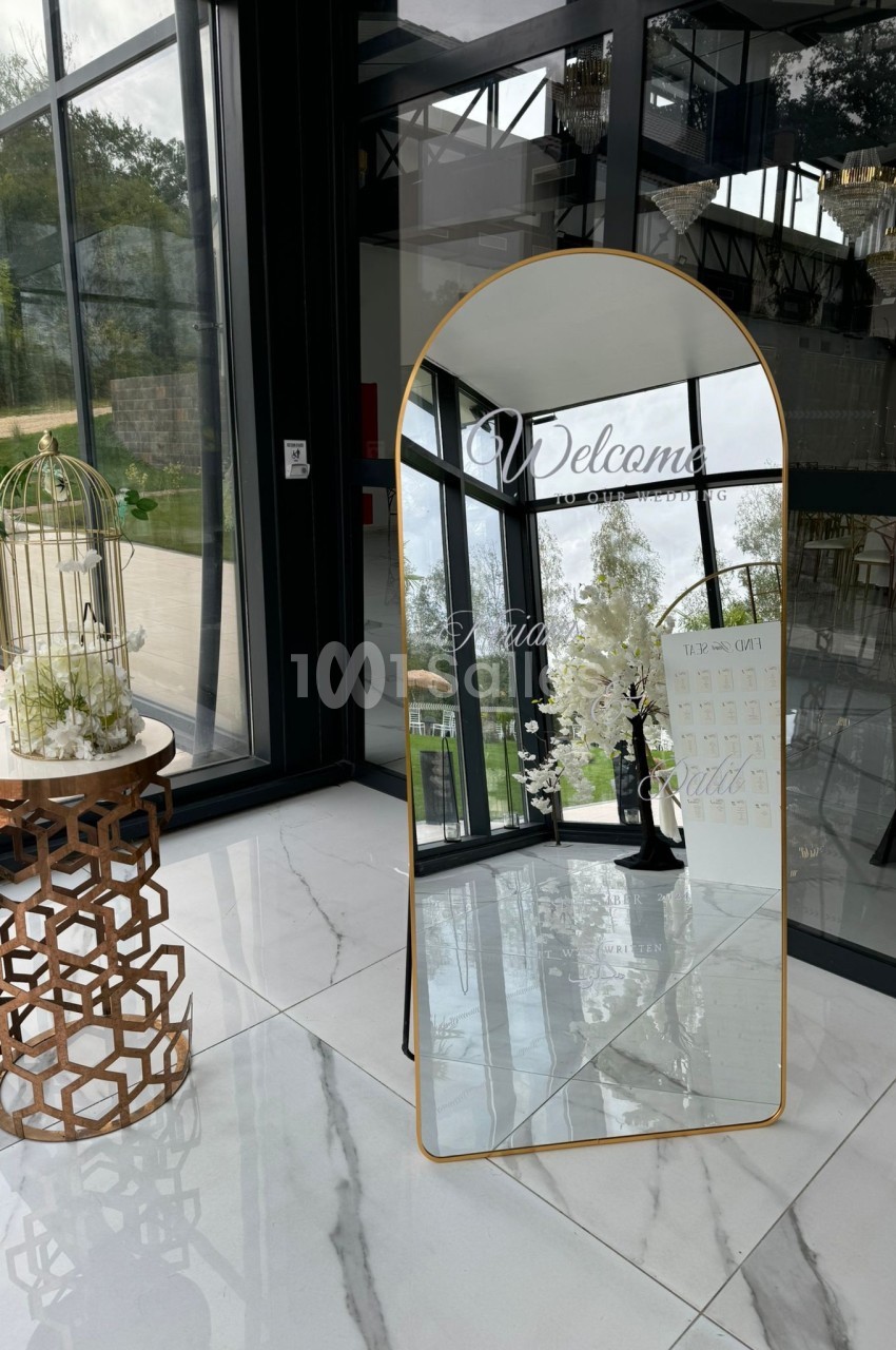Grand miroir doré avec inscription de bienvenue, placé dans un espace lumineux près d'une table décorative et d'une cage.