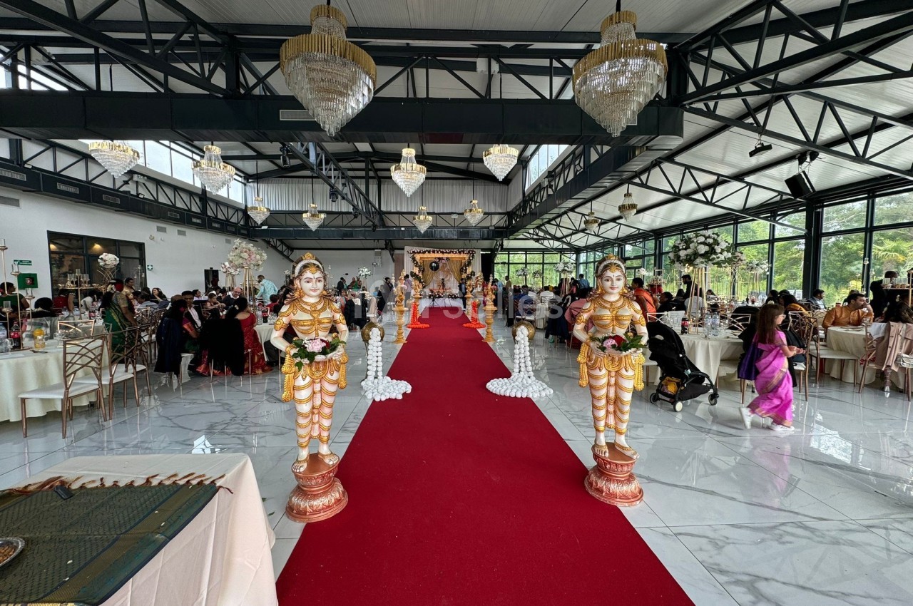 Salle de réception décorée pour un mariage avec tapis rouge, statues traditionnelles et invités assis à des tables.