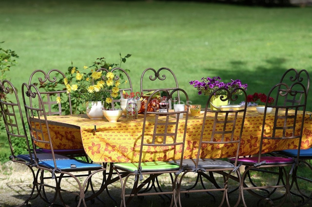 Table de jardin avec nappe jaune décorée, entourée de chaises en fer forgé, ornée de fleurs et de vaisselle.