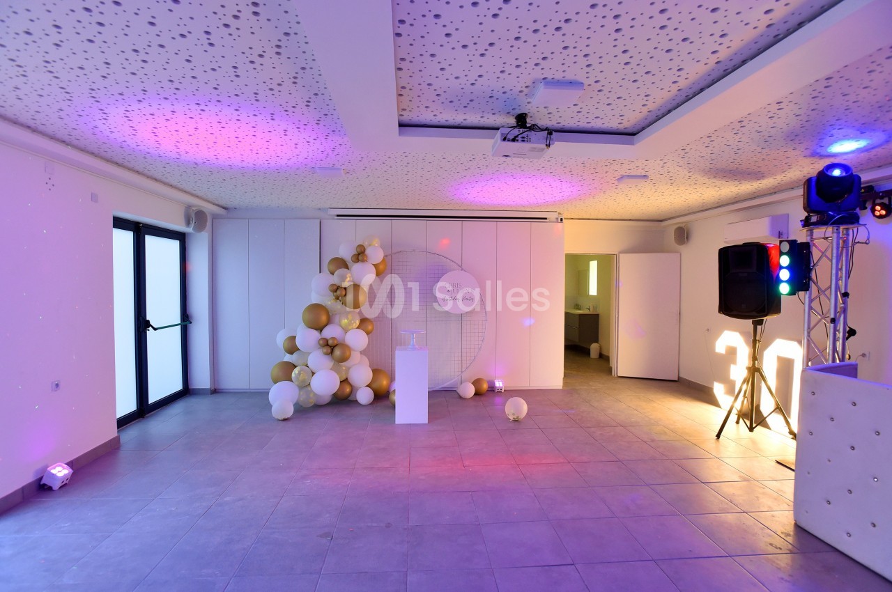Salle décorée avec des ballons blancs et dorés, éclairage coloré, sol en carrelage et plafond perforé.