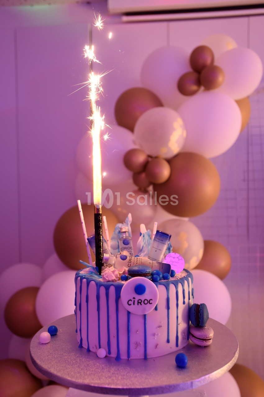 Gâteau décoré avec des bouteilles miniatures, une fontaine lumineuse et des ballons blancs et dorés en arrière-plan.
