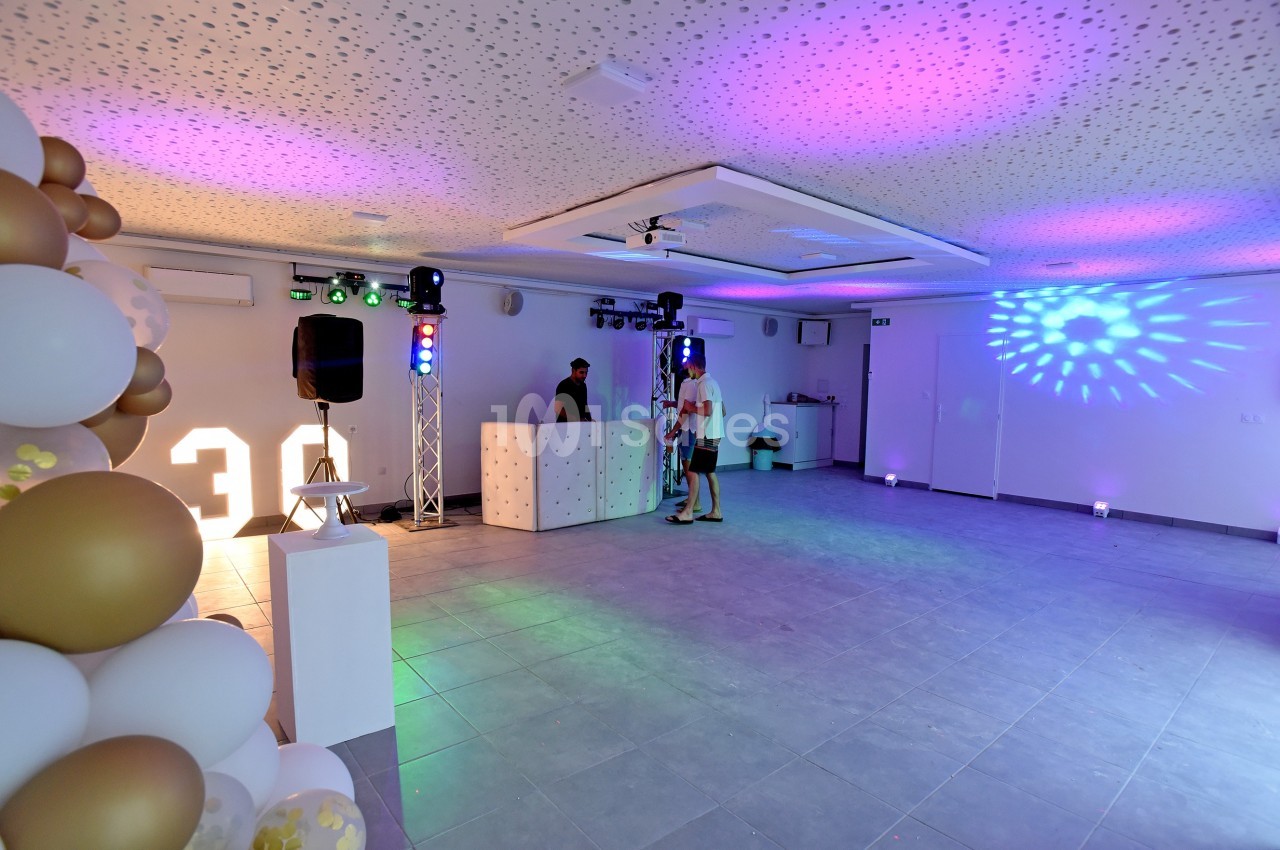 Salle décorée avec des ballons, éclairages colorés et une table de DJ, prête pour un événement festif.