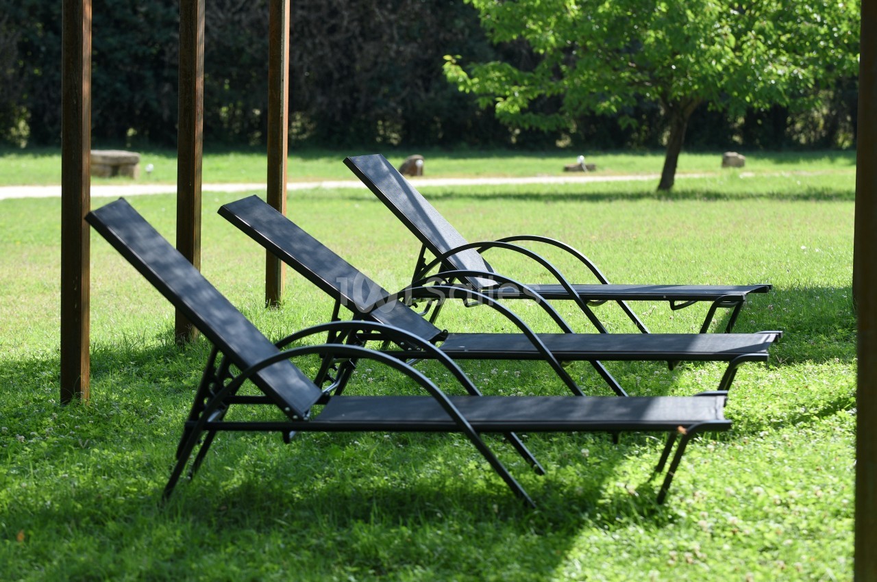 Trois chaises longues noires alignées sur une pelouse verte sous une structure en bois, avec des arbres en arrière-plan.