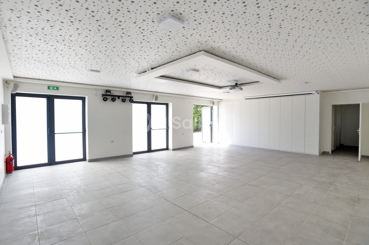Salle vide avec carrelage gris, plafond acoustique perforé, grandes baies vitrées et porte ouverte sur l'extérieur.
