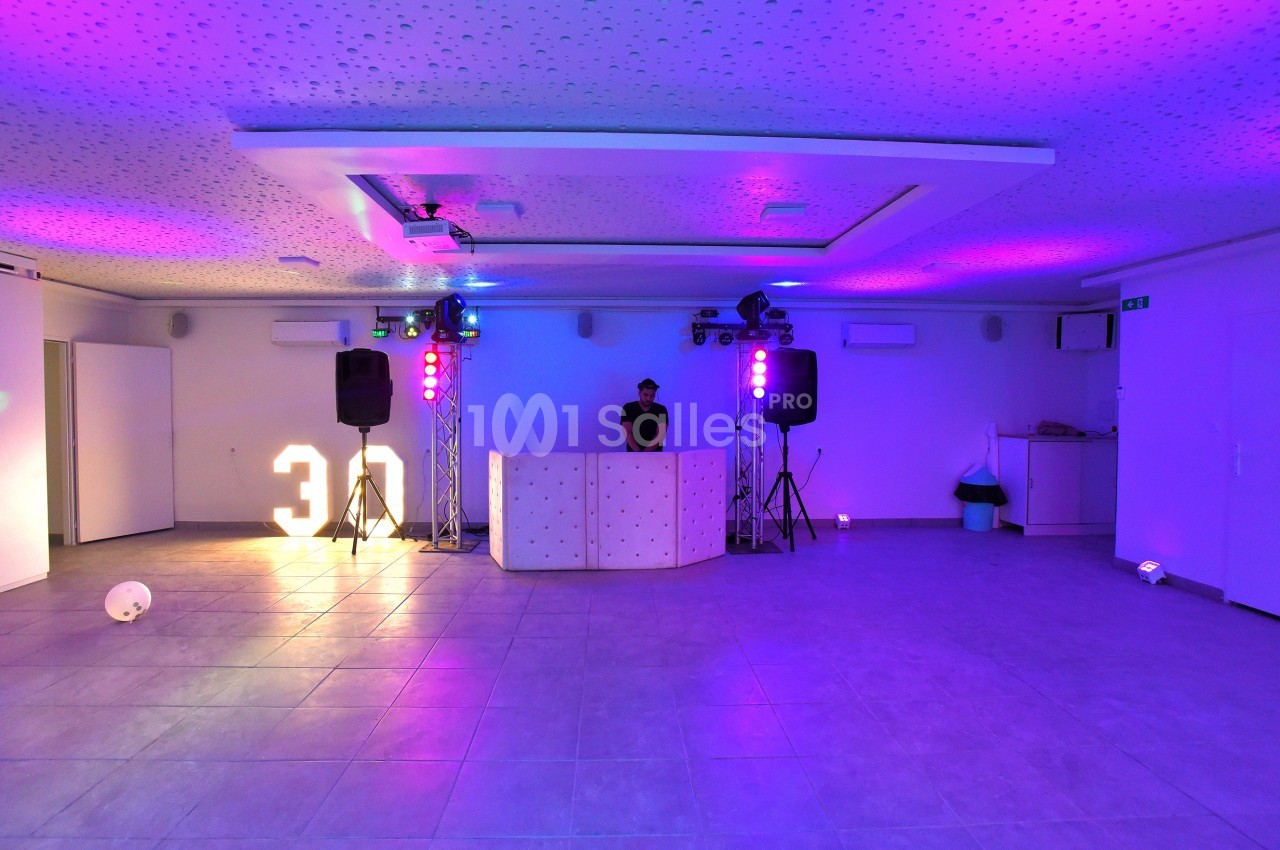 Salle éclairée par des lumières colorées avec un DJ derrière une table, décorée pour un événement festif.